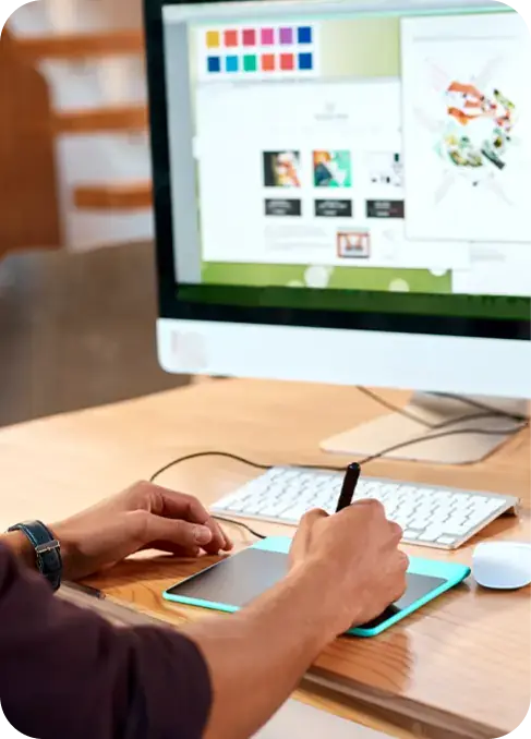 Person using a digital drawing tablet with a stylus, working on graphic design software displayed on a computer monitor. The screen shows various color swatches and design elements.